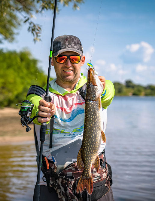 Angler holding a spinning rod and showcasing a caught pike near the riverbank, wearing fishing gear and Select-branded clothing.
