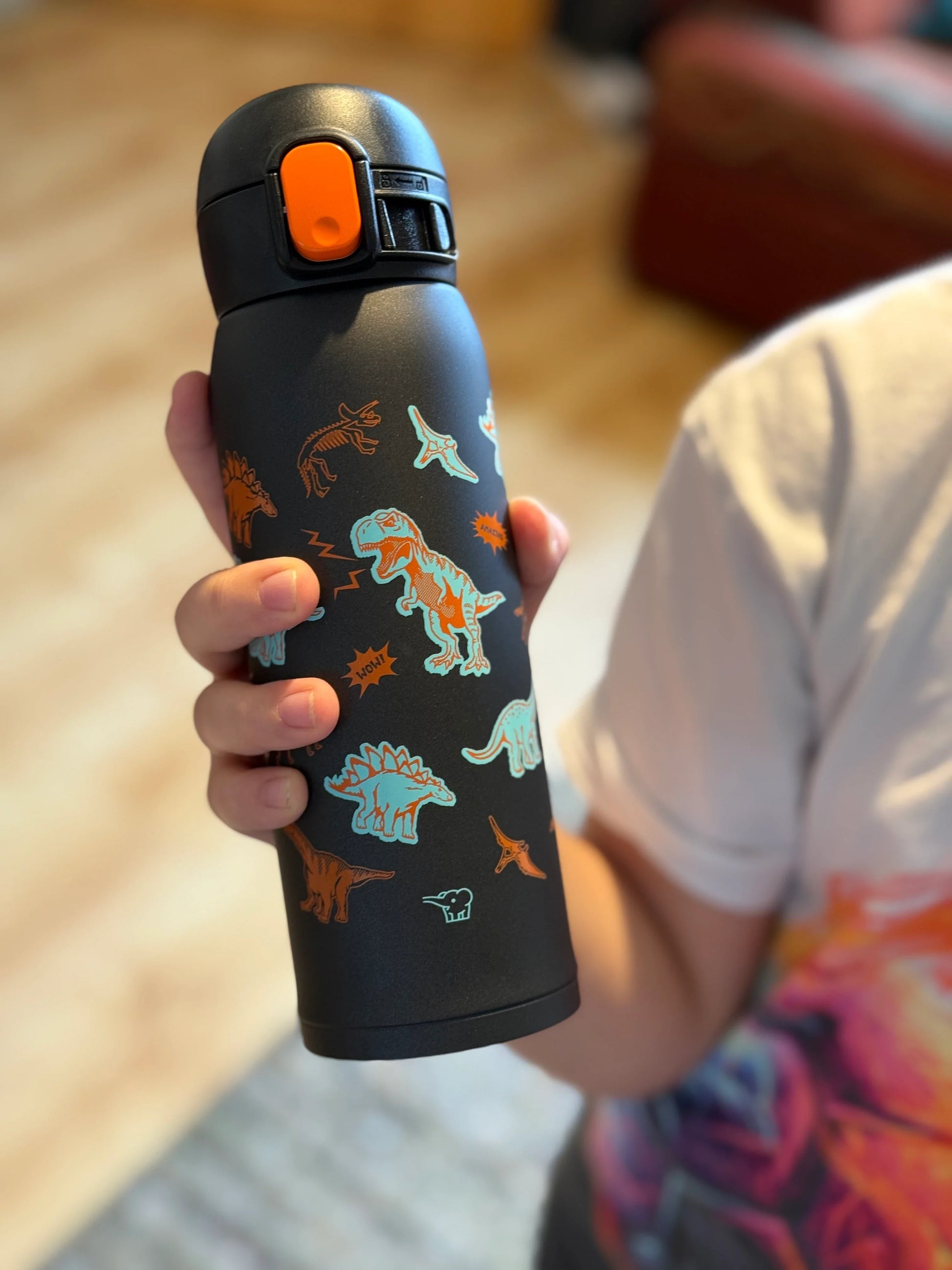 Zojirushi SM-WM48BA kids thermal mug 480 ml (16.20 oz) in black with colourful dinosaur design, reusable insulated travel bottle for hot and cold drinks, held in childโs hand indoors.